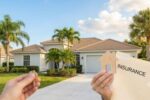 A hand holding a wooden house cutout and an "Insurance" folder in the foreground, with a sunlit, modern Florida home and palm trees in the background.