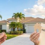 A hand holding a wooden house cutout and an "Insurance" folder in the foreground, with a sunlit, modern Florida home and palm trees in the background.