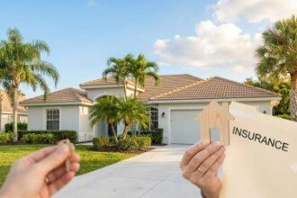 A hand holding a wooden house cutout and an "Insurance" folder in the foreground, with a sunlit, modern Florida home and palm trees in the background.