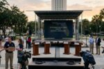 An elevated photo of an empty outdoor stage at sunset in front of the Florida State Capitol, with a large LED screen that reads "FLORIDA STATEWIDE RACES 2026: DEBATE STATUS CANCELLED." Empty podiums and media equipment are in the foreground, surrounded by people holding protest signs.