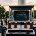 An elevated photo of an empty outdoor stage at sunset in front of the Florida State Capitol, with a large LED screen that reads "FLORIDA STATEWIDE RACES 2026: DEBATE STATUS CANCELLED." Empty podiums and media equipment are in the foreground, surrounded by people holding protest signs.